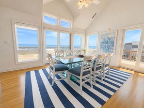 Un comedor con mesa de vidrio y sillas blancas, ventanas amplias con vistas al océano y un tapete a rayas azules.