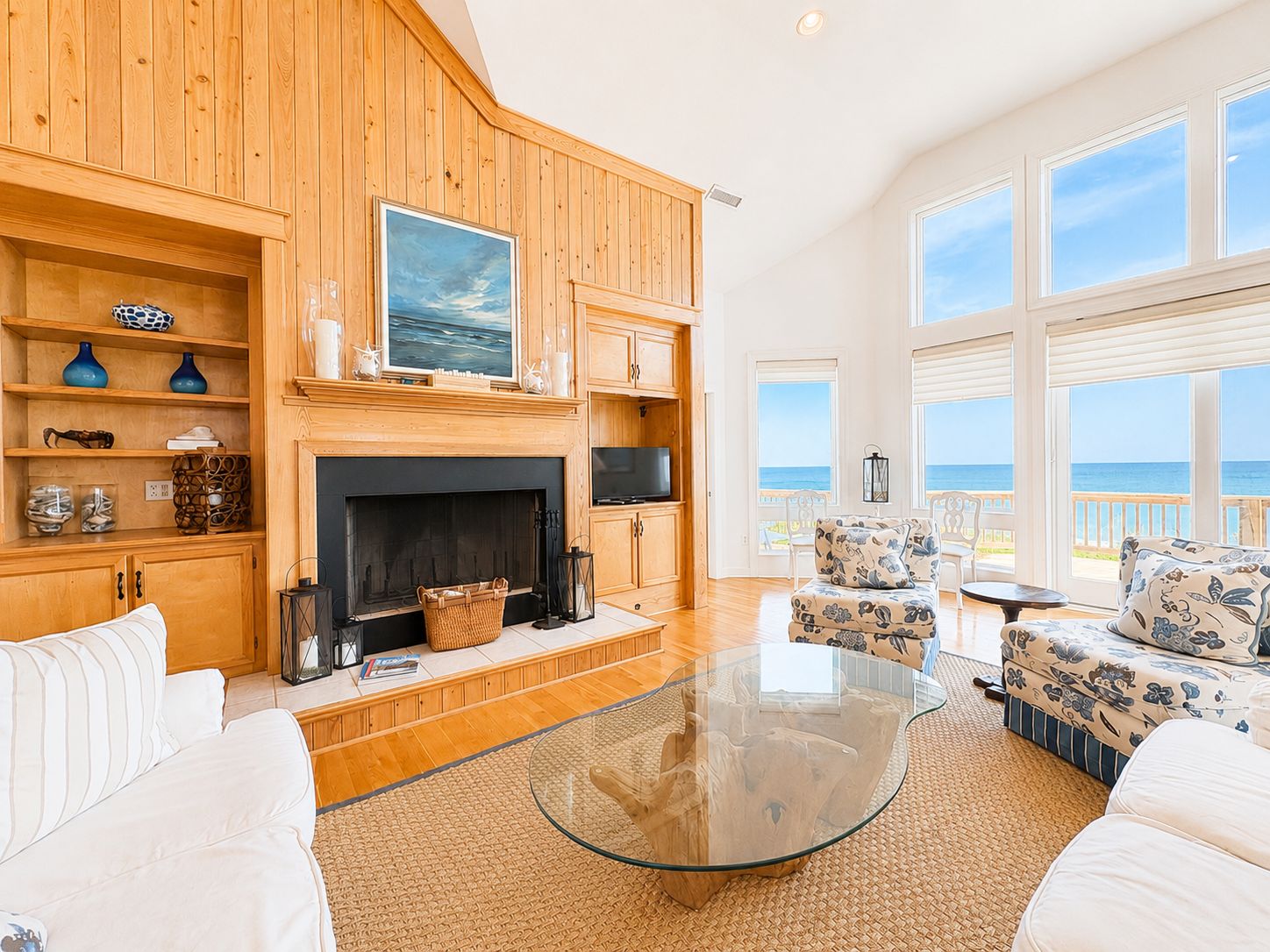 A spacious living room with a fireplace, wooden shelves, and a large window overlooking the sea.