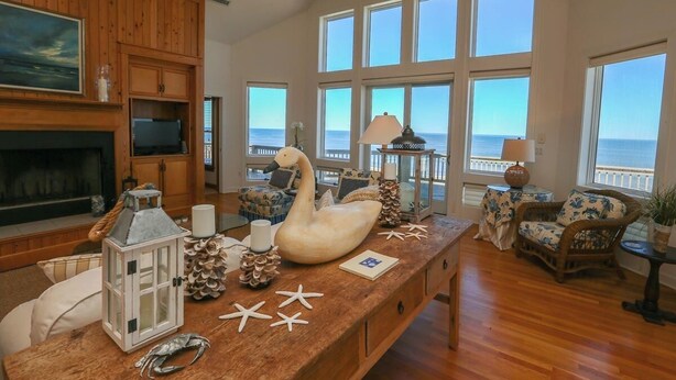 Un salón espacioso con suelo de madera, ventanas grandes con vistas al mar, chimenea y un centro de mesa con forma de cisne.