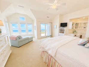 A bedroom with a large bed, a desk, a television, and a view of the sea through the windows.