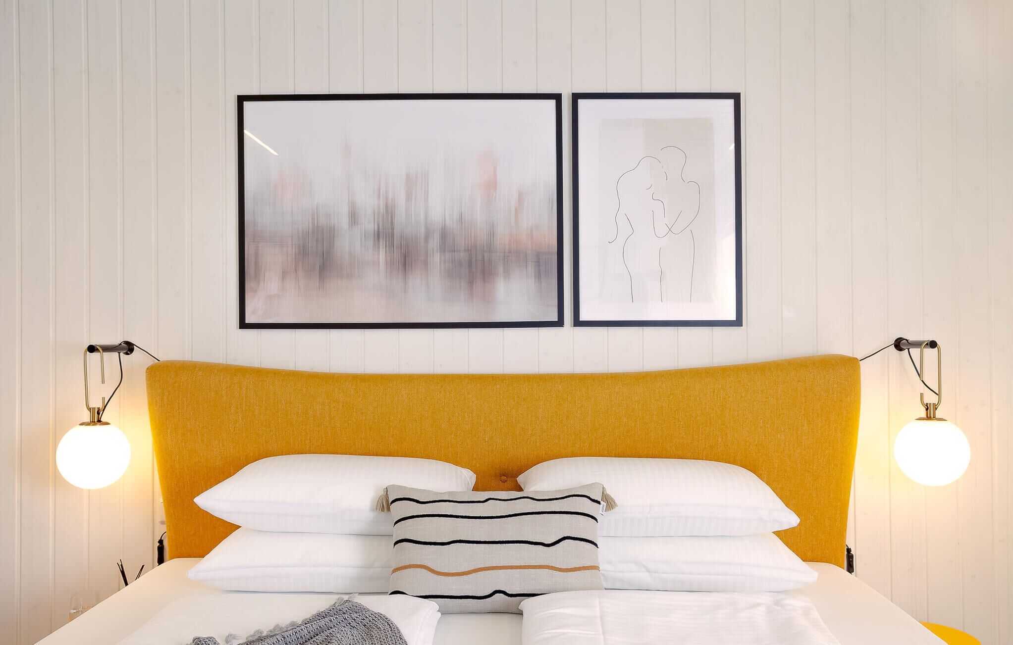 A bed with a yellow headboard, white bedding, and striped pillows. There are two framed artworks above the bed.