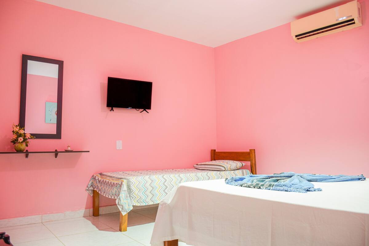 Un dormitorio con paredes rosadas, un televisor montado en la pared, una cama con sábana blanca y una mesita de noche con un jarrón de flores.