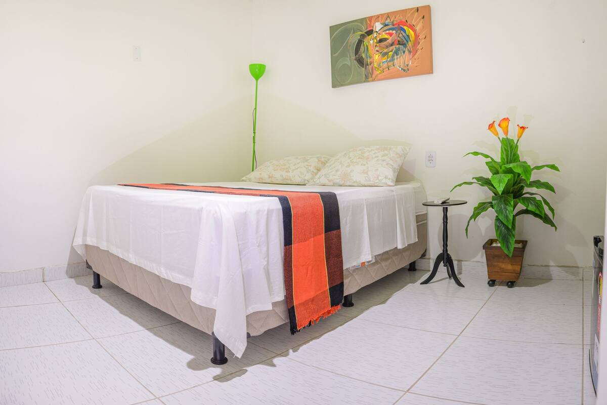 Un dormitorio con cama, mesita de noche, lámpara de pie verde, un cuadro en la pared y una planta en maceta.