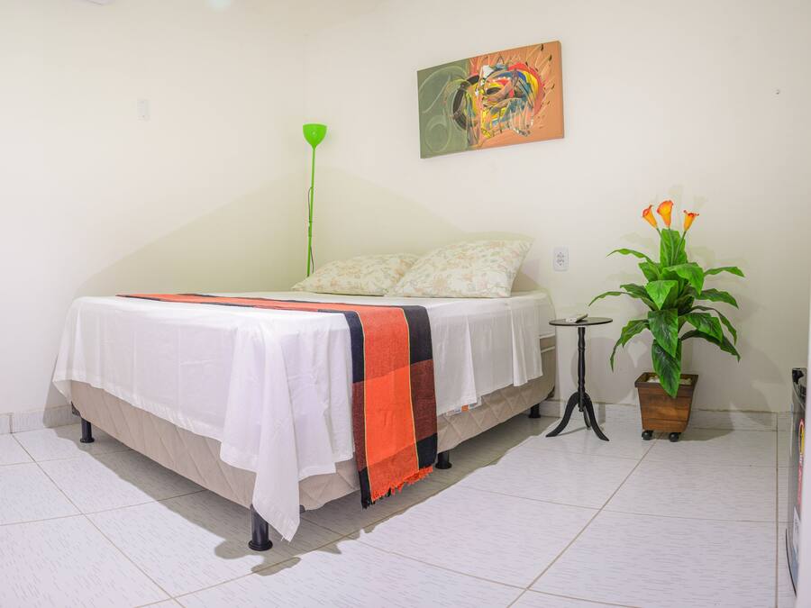 Quarto com cama, mesa de cabeceira, luminária de chão verde, um quadro na parede e planta em vaso.