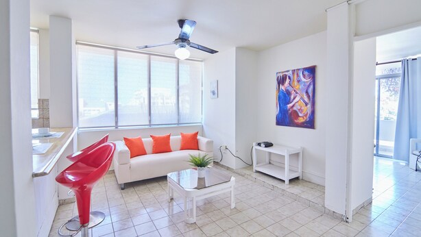 A modern living room with a white sofa, red chair, glass coffee table, and a ceiling fan. - Rentamos Aparta Estudio for Short or lon (Santo Domingo)
