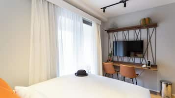 Una habitación de hotel moderna con una cama grande, un escritorio con dos sillas, un televisor montado en la pared y un ventanal con cortinas.