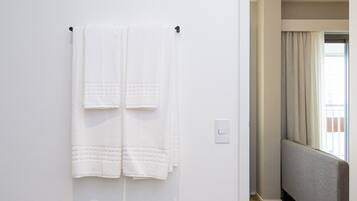 Habitació d'hotel amb una paret blanca, un penjador amb dues tovelloles, un terra de fusta i vista a una altra habitació per una porta oberta.