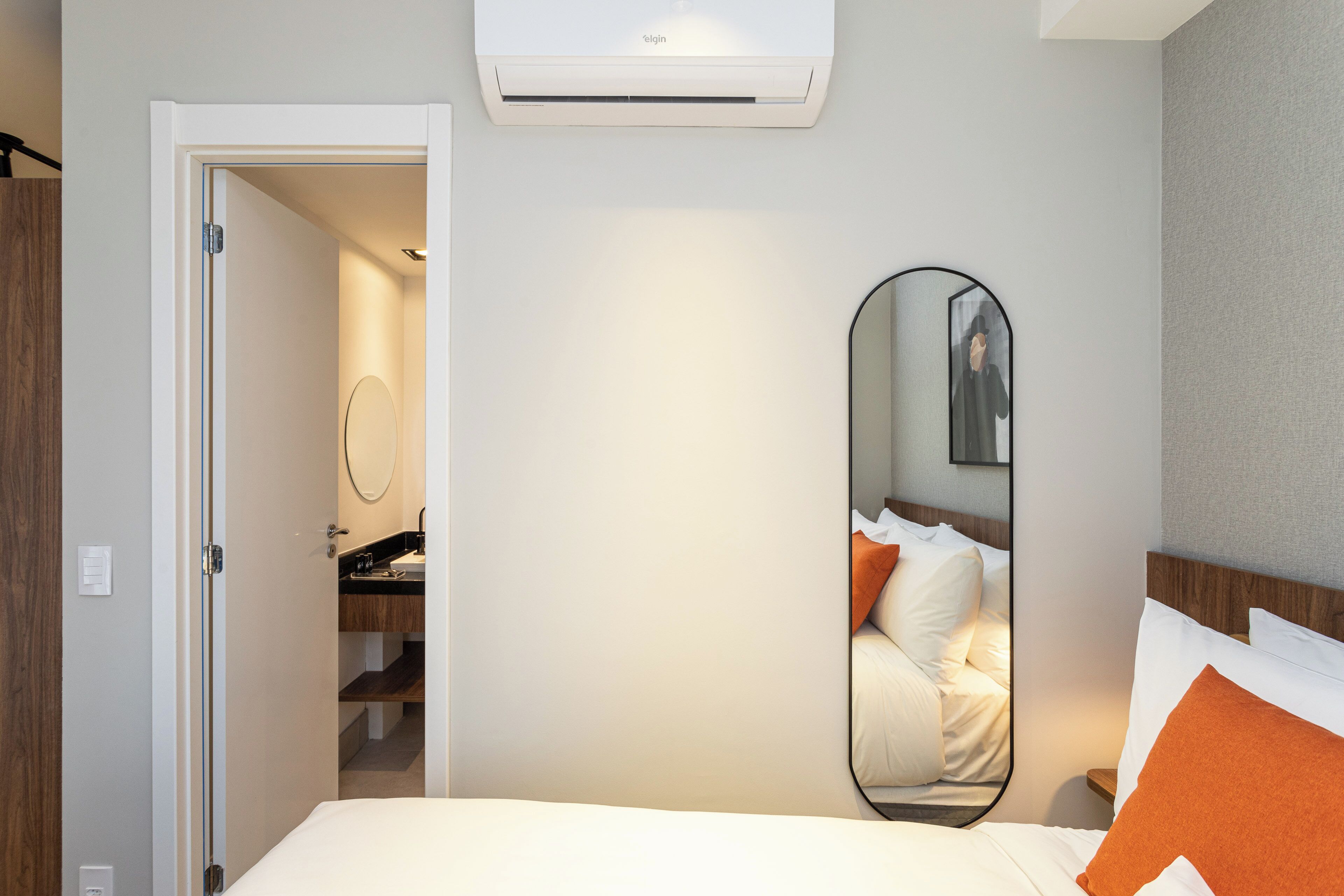 A hotel room with a bed, a bedside mirror, a wardrobe, and an air conditioning unit.