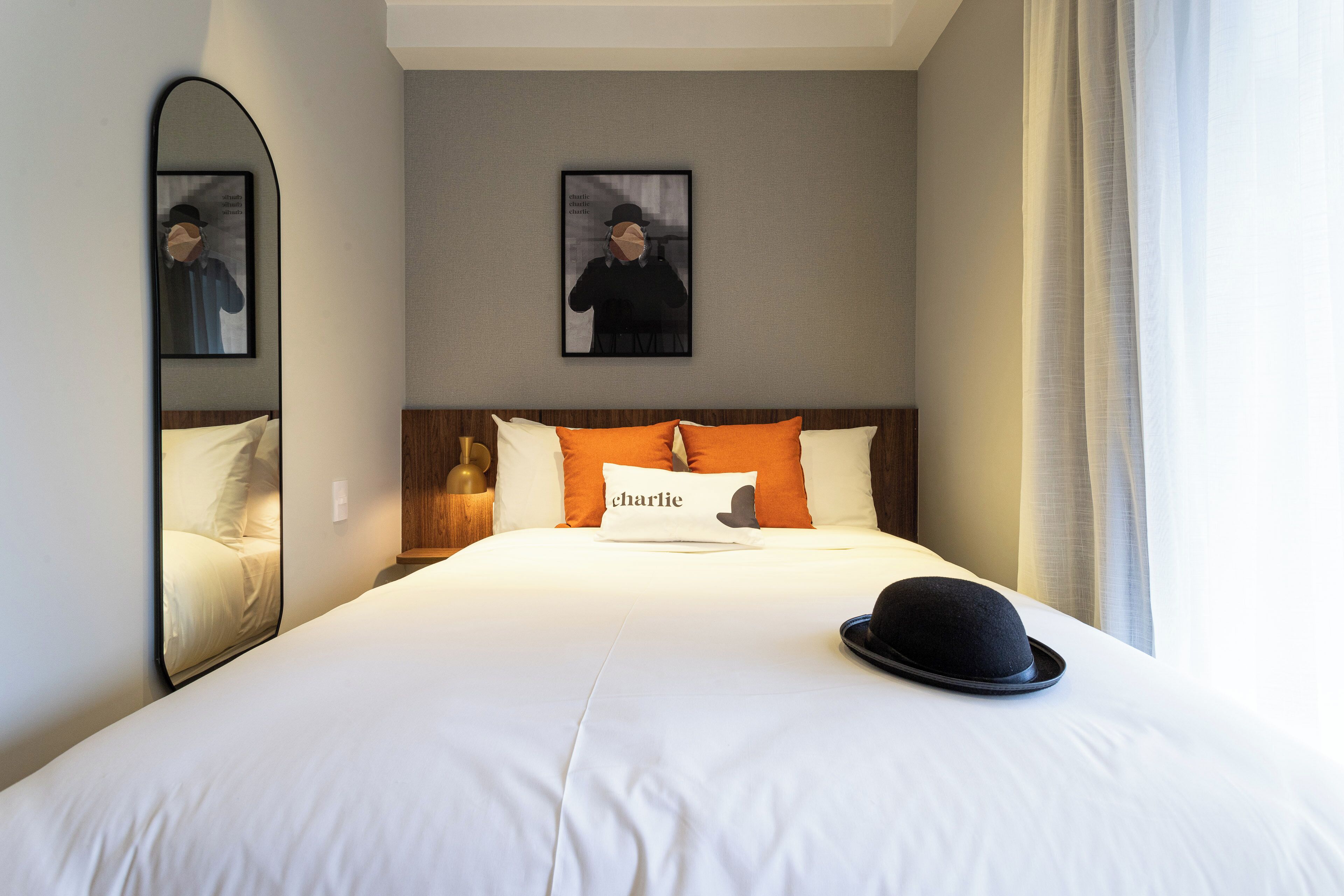A neatly made bed with white linens, a pillow with the word "charlie," a black hat on the bed, and a framed picture on the wall.