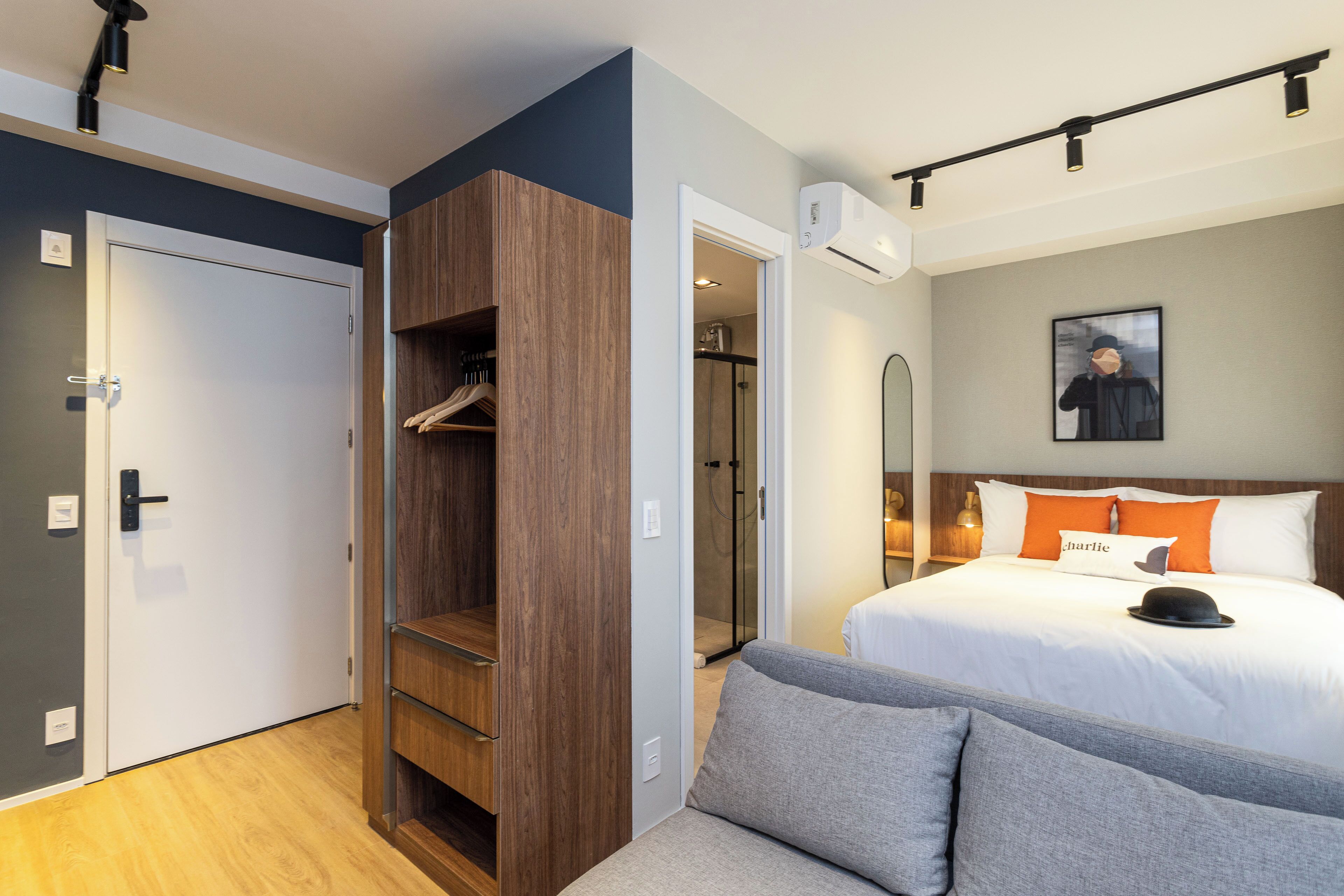A modern hotel room with a wardrobe, a bed with white bedding and orange pillows, a grey sofa, and a framed picture on the wall.