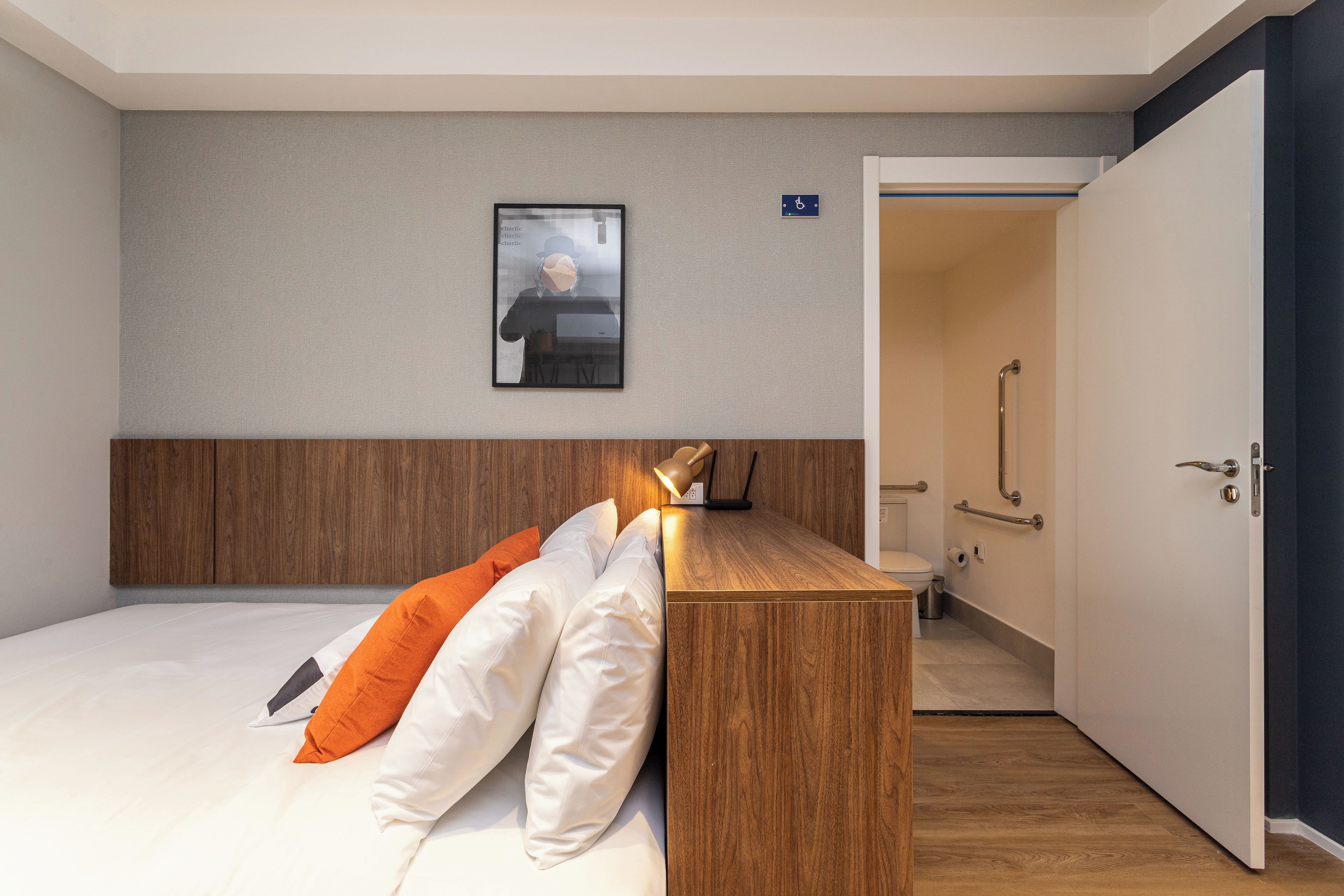 A hotel room with a bed, wooden headboard, white and orange pillows, a small desk with a lamp, a framed picture on the wall, and a door leading to a bathroom.