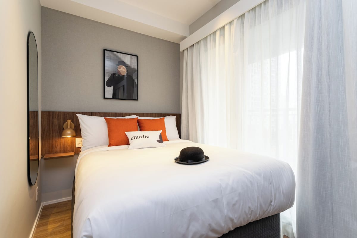 A neatly made bed with white bedding and orange pillows, a black hat on the bed, a framed picture on the wall, and a window with sheer curtains.