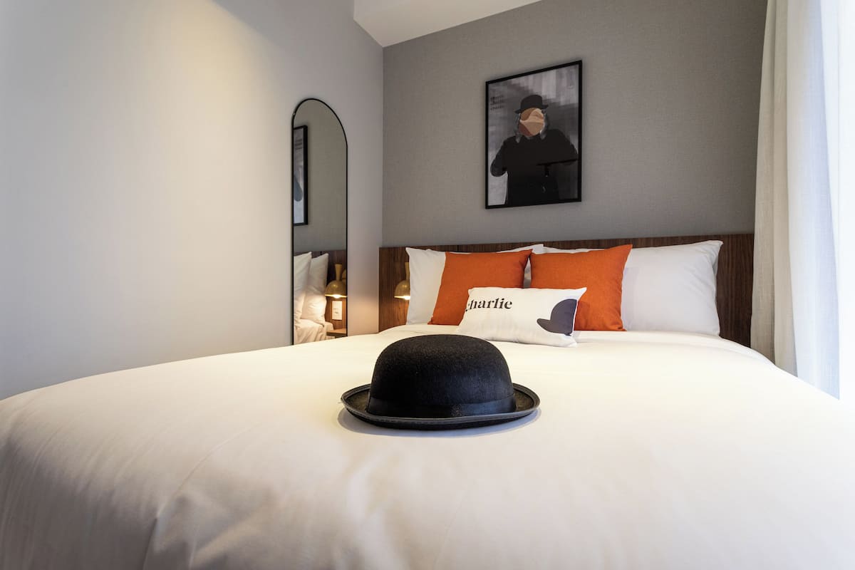 A neatly made bed with a black hat on top, a pillow with the word "Charlie," and a framed picture on the wall.