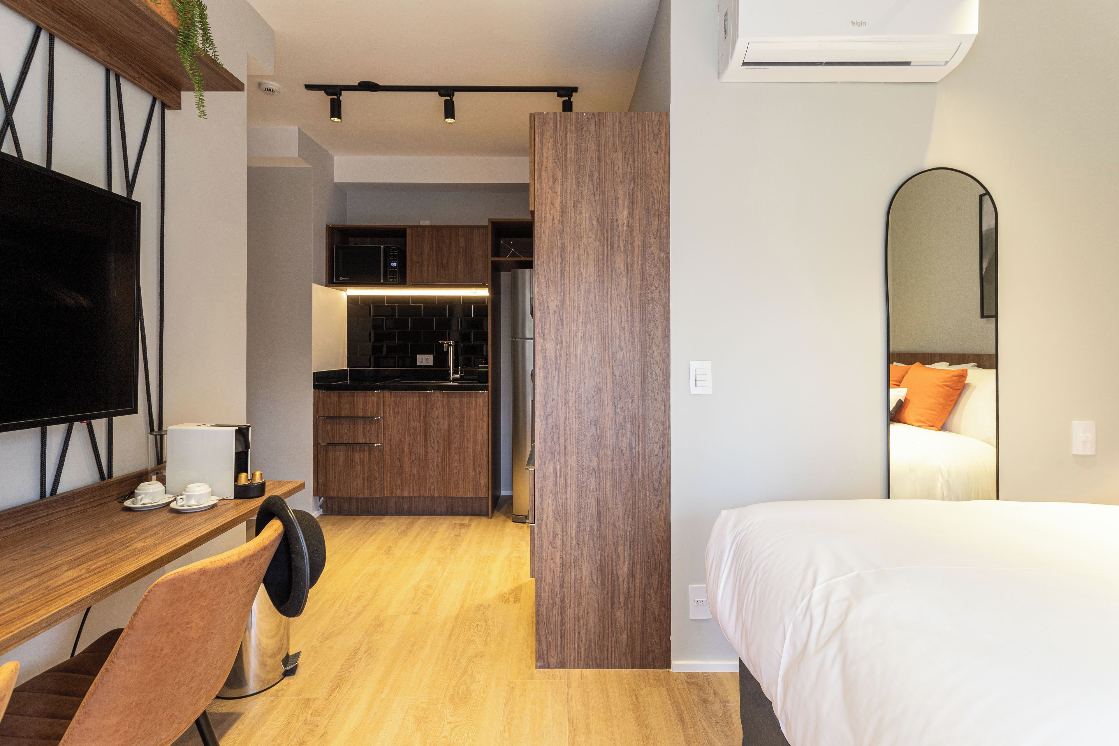 A modern hotel room with a wooden desk, a chair, a bed, and a kitchen area in the background.