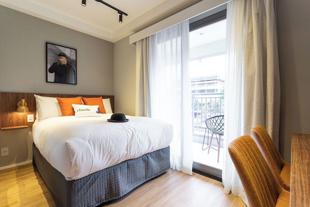 A modern hotel room with a large bed, a small table, a chair, and a balcony with a view.