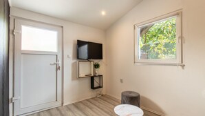 A room with a white door, a mounted TV, a small table, a window with a view of greenery, and a wooden floor.