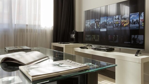 Una sala de estar moderna con un televisor de pantalla plana grande que muestra carteles de películas, una mesa de centro de vidrio con revistas y un mueble blanco elegante.