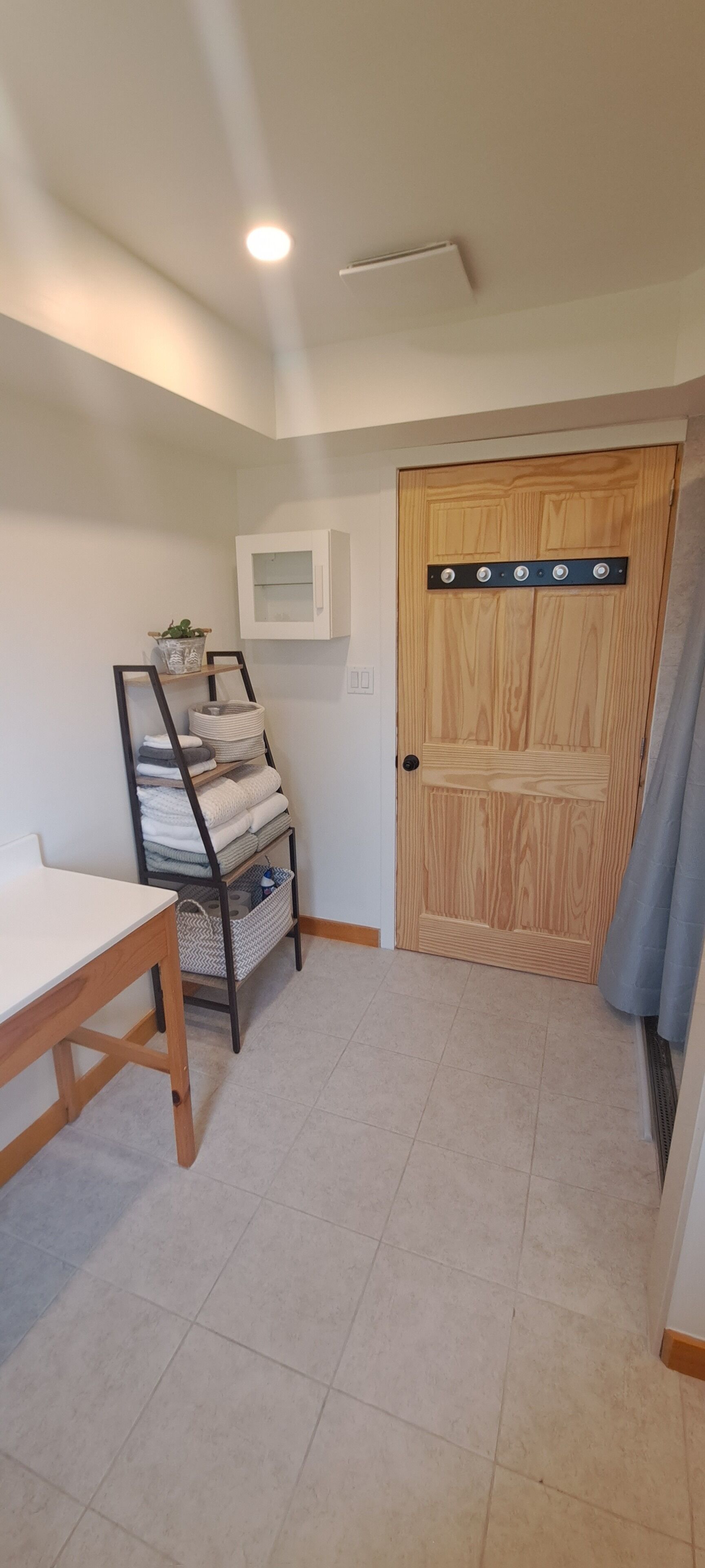 Une petite pièce bien rangée, avec une porte en bois, une armoire blanche, un présentoir en métal où sont pli ées des serviettes et une table en bois.