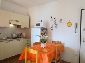Une cuisine compacte avec des meubles blancs, un four à micro-ondes et un réfrigérateur. Une table à manger est dressée, recouverte d’une nappe orange et entourée de chaises en bois.