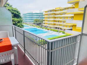 Un balcon avec une piscine, des chaises blanches et une table sur laquelle se trouve un panier de fruits.