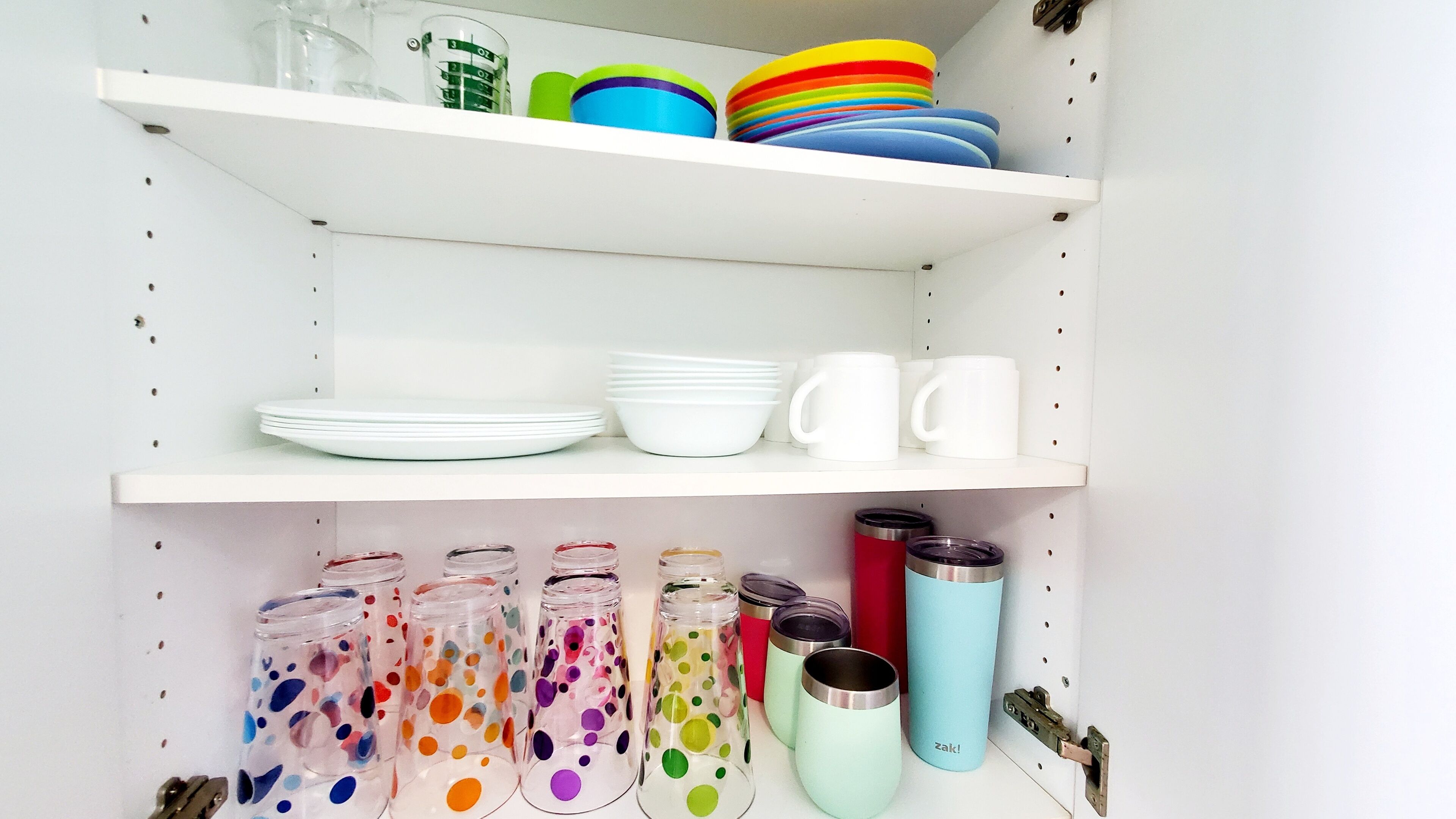 A kitchen shelf with white plates, bowls, and mugs, and colorful glass jars with lids.