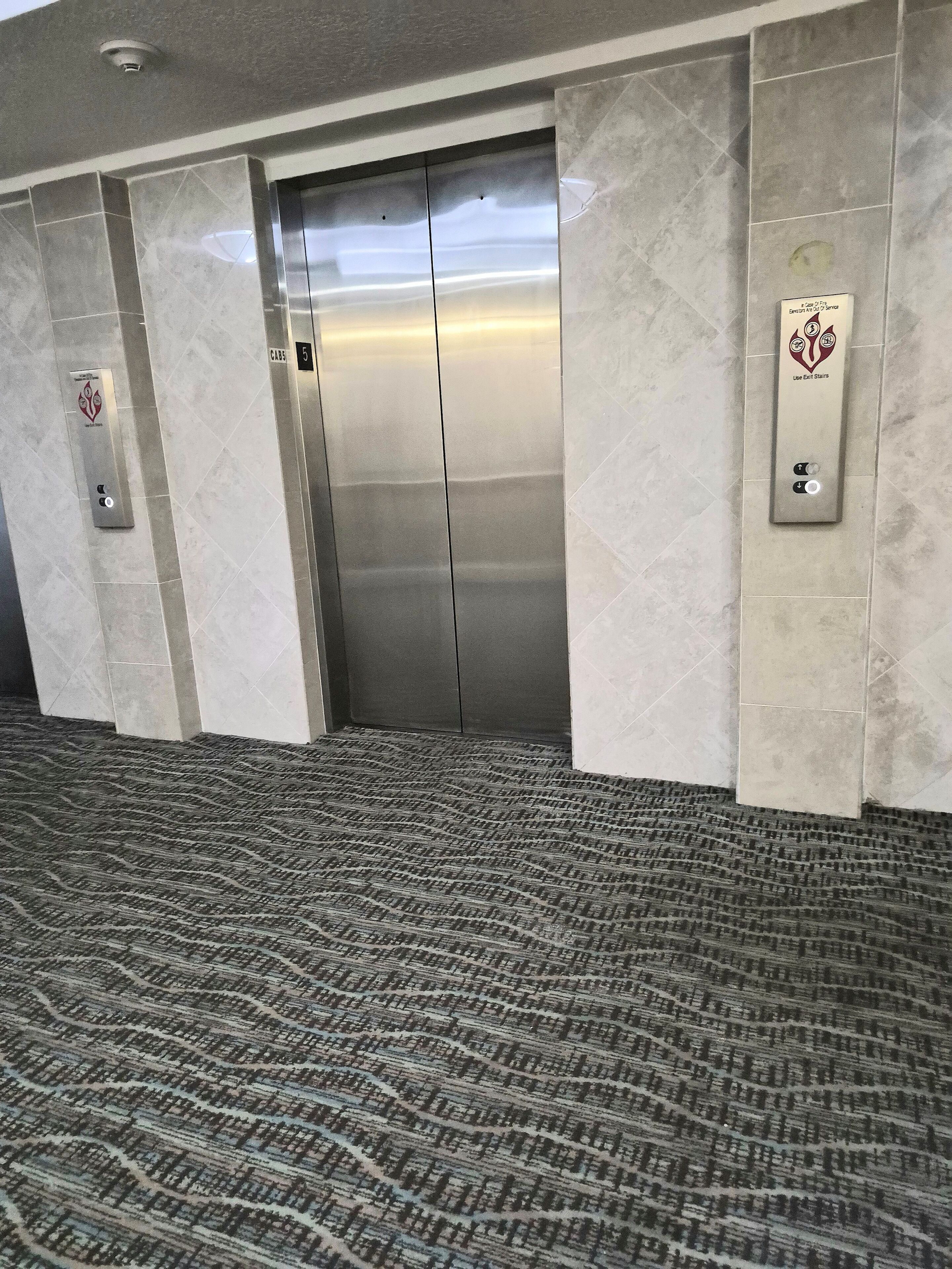 Un ascenseur aux finitions métalliques, un sol recouvert de moquette et un panneau mural avec un logo.