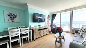 Een woonkamer met een televisie, een eettafel, stoelen, een bank en uitzicht op zee.