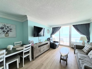 Uma sala de estar com televisão, mesa de jantar, cadeiras, sofá e varanda com vista.