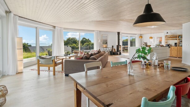 Amplia sala de estar con mesa de comedor de madera, sofá y vistas al exterior a través de grandes ventanales.