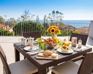 Une table de petit-déjeuner dressée sur un balcon avec vue sur la mer, comprenant des assiettes, des tasses et un vase rempli de tournesols.
