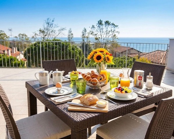 Une table de petit-déjeuner dressée sur un balcon avec vue sur la mer, comprenant des assiettes, des tasses et un vase rempli de tournesols.