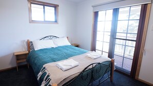 A neatly made bed with a teal bedspread and white pillows, a bedside table with a lamp, and a window with blinds.