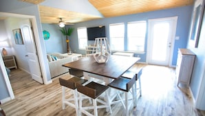A spacious dining area with a wooden table and white chairs, a ceiling fan, and large windows allowing natural light. - NEW: Beach House 3-Bed/2-Bath with Ocean View & Golf Cart -- Great Beach (Surfside Beach)