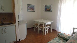 A compact kitchen with a sink, a white refrigerator, a dining table with four chairs, and two framed pictures on the wall. - Seaside Welcoming Flat Next to the Beach - Beahost (San Michele al Tagliamento)