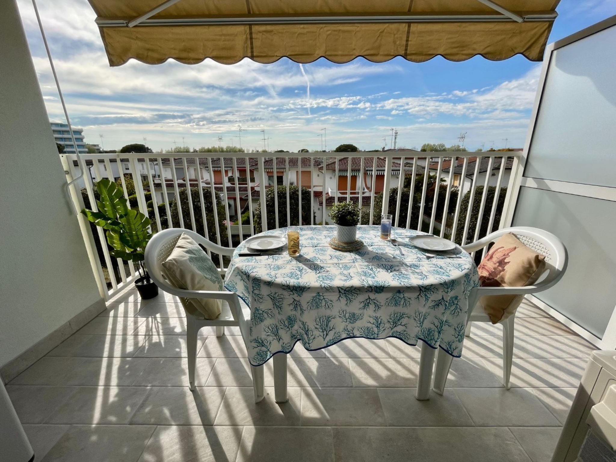 Een balkon met een tafel gedekt voor twee personen, stoelen, een plant in pot en gebouwen op de achtergrond.