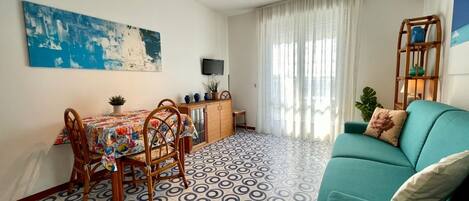 A living room with a teal sofa, a dining area with a table and chairs, a TV mounted on the wall, and a patterned floor.
