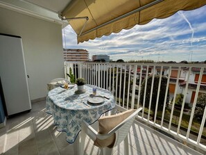 A balcony with a table set for two, a potted plant, and a view of buildings in the background. - Comfy Studio With Shared Pool (Caorle)