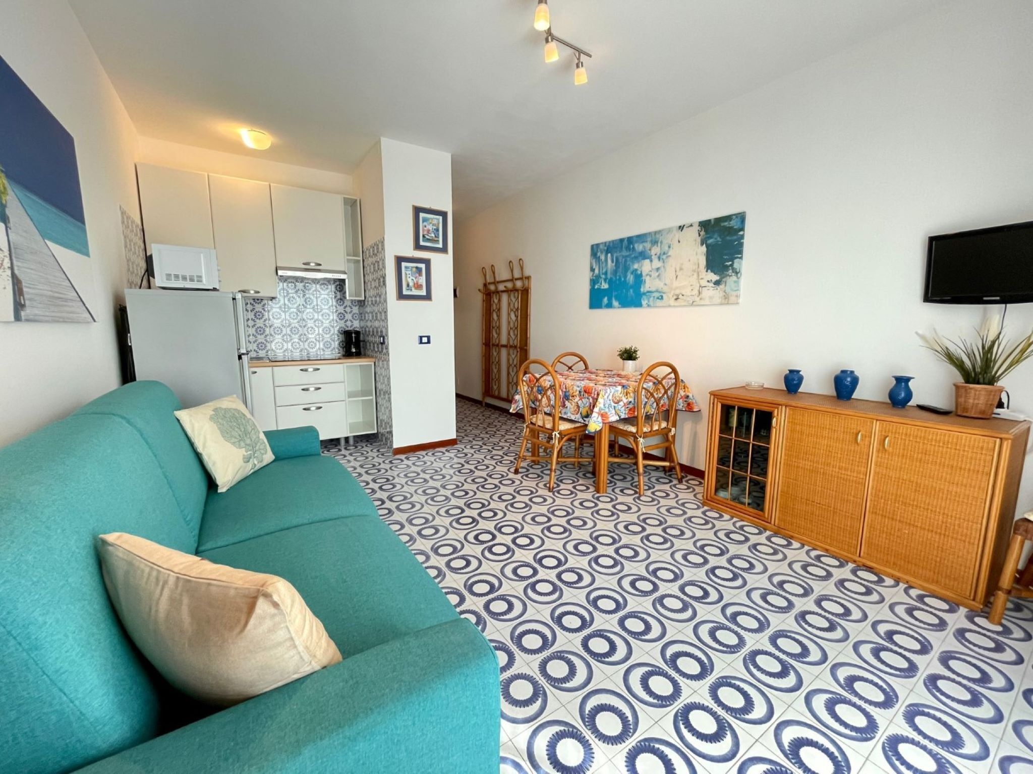 Een knus woongedeelte met een blauwgroene bank, een eethoek met tafel en stoelen, een kitchenette en een televisie die aan de muur hangt.