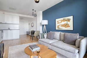 Een moderne woonkamer met een grijze bank, een salontafel met boeken, een eethoek met een tafel en stoelen, en een keuken met witte kasten.