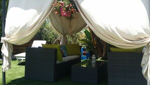 A covered outdoor seating area with wicker furniture and cushions, surrounded by greenery. - Gabriel Villa Cyprus (Paralimni)
