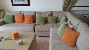 A living room with a beige sofa, green and orange cushions, a glass coffee table with remote controls and a candle, and a framed picture on the wall. - Gabriel Villa Cyprus (Paralimni)