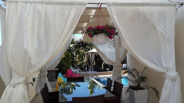 A covered outdoor seating area with a pool, white curtains, and hanging plants. - Gabriel Villa Cyprus (Paralimni)