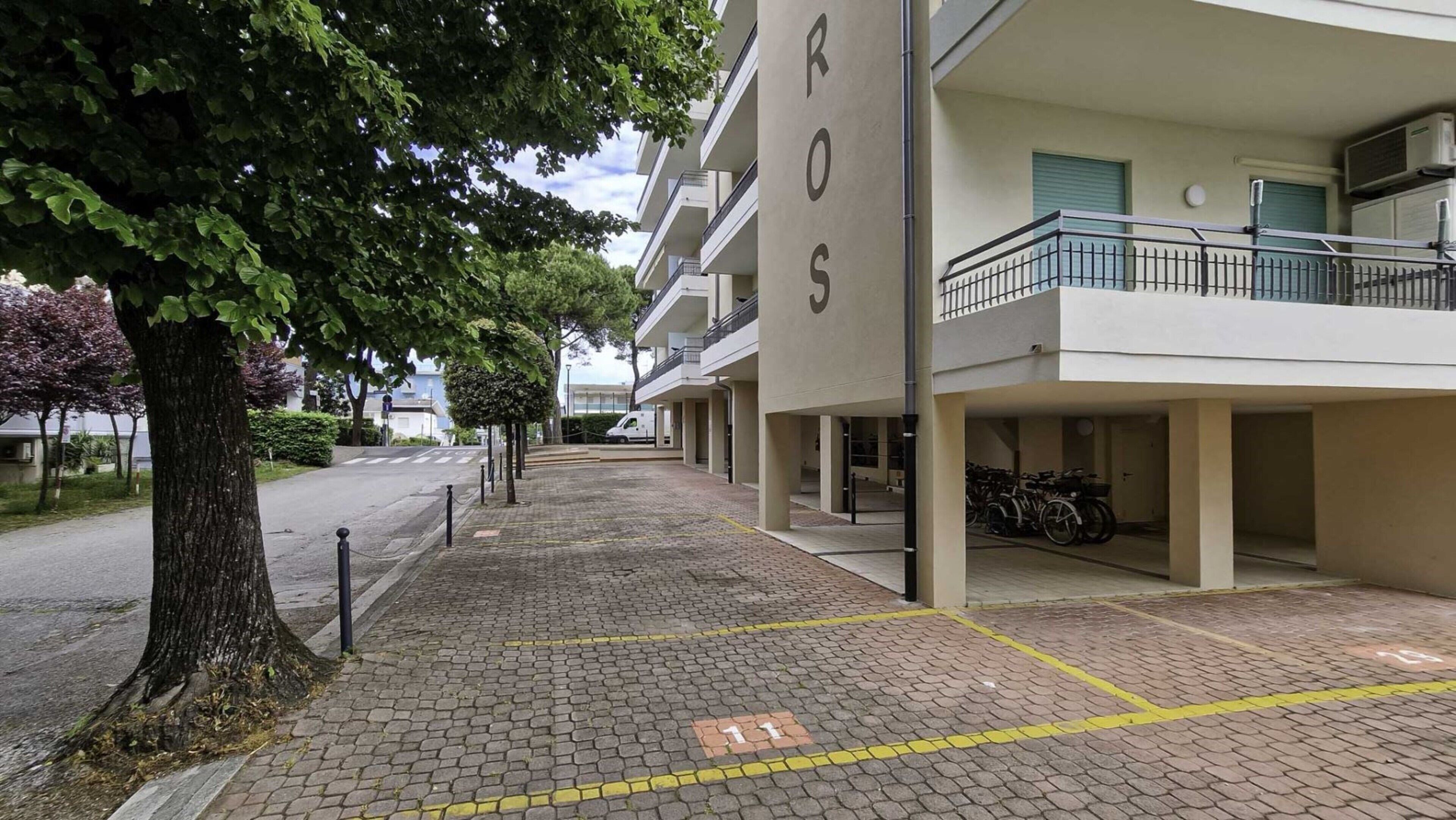 Ein Gebäude mit der Aufschrift 'ROS' an der Fassade, ein überdachter Parkbereich mit Fahrrädern und ein von Bäumen gesäumter Bürgersteig.