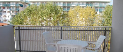 A balcony with white plastic chairs and a table, overlooking a building with balconies and greenery.