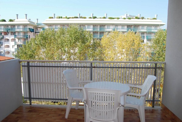 Uma varanda com cadeiras e mesa de plástico branco, com vista para um edifício com varandas e vegetação.