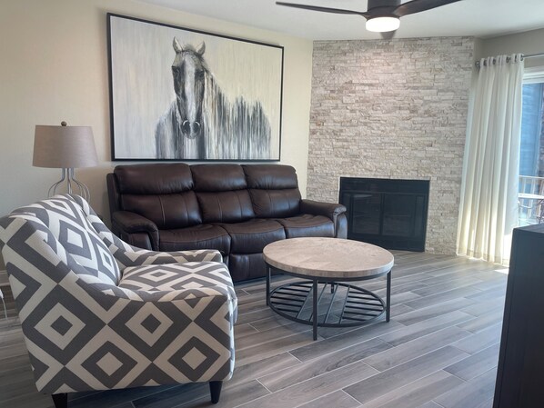 A living room with a brown leather sofa, a patterned armchair, a round coffee table, a fireplace, and a large abstract painting of a horse.