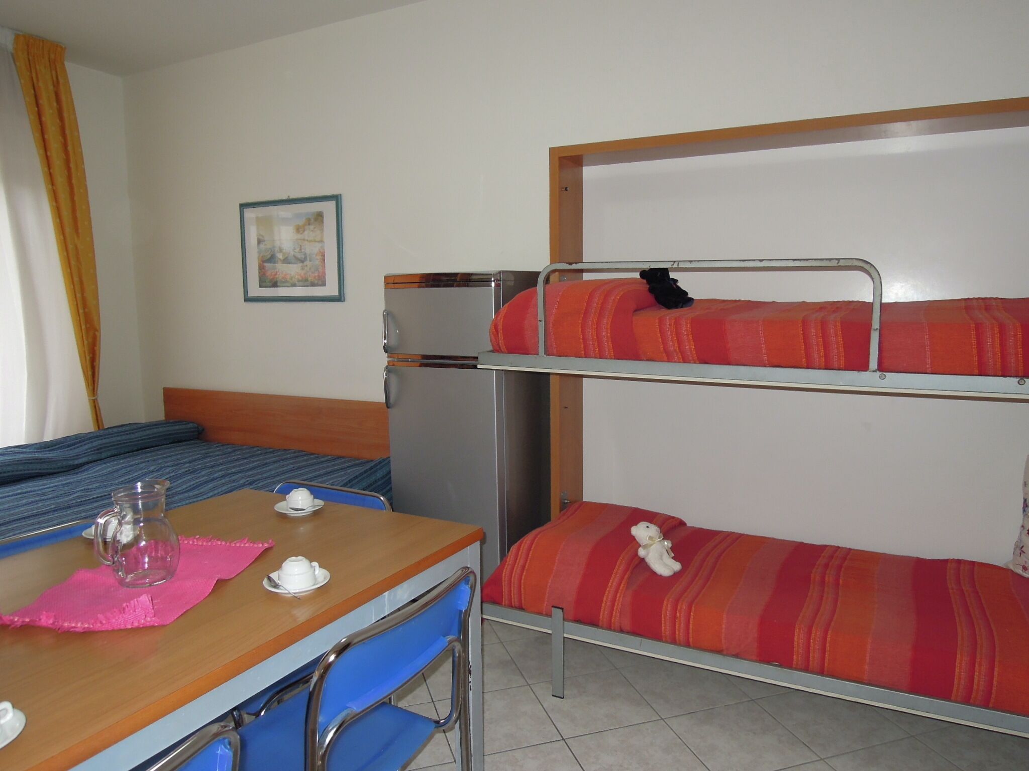 Ein Etagenbett mit zwei Schlafstellen, ein Holztisch mit Stühlen, ein kleiner Kühlschrank und ein Bild an der Wand.