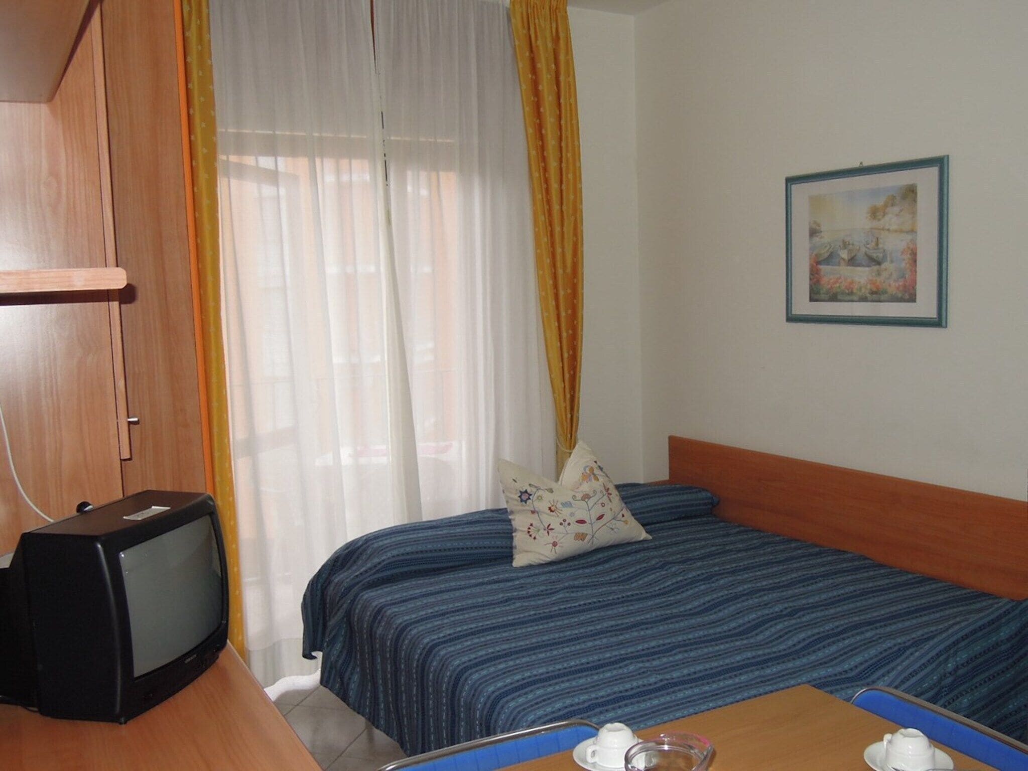 A hotel room with a bed, a television, a painting on the wall, and a window with curtains.