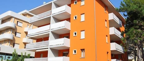 A multi-story building with balconies, a parking area, and greenery.