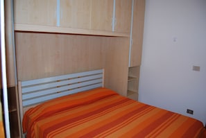 Yhden hengen sänky, jossa on raidallinen peitto, puinen pääty ja sen yläpuolella oleva puinen vaatekaappi.